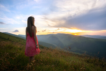 Young happy woman traveler in red dress standing on grassy hillside on a windy evening in summer mountains enjoying view of nature at sunset