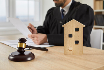 Fototapeta na wymiar Close up of a gavel and a small symbolic toy house on a wooden table in court, with a judge working with settlement documents in the background. Real estate law, property, foreclosure concepts