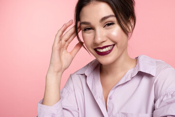 Laughing modern young caucasian girl with perfect trendy make up, glowing healthy skin wearing gentle lilac shirt having fun on pink studio background. Positive emotions, skincare products advertising