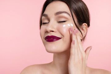 Pleased young woman with bare shoulders and red lips applying daily face moisturizer, smiling girl use facial moisturizing cream on pink studio background, enjoying skin condition.