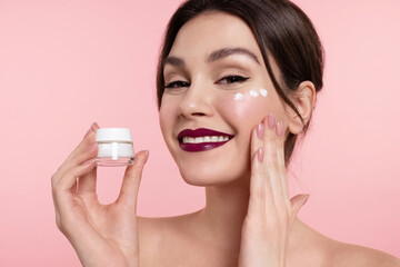 Pretty young woman applying moisturizing facial cream on cheek, holding template jar of skincare cosmetics product, looking at camera. Happy girl with bare shoulders enjoying beauty spa procedure.