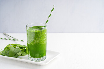 Green healthy homemade vegan smoothie made of raw spinach leaves with high nutritional value and source of vitamins served in drinking glass with paper straw on white wooden table with copy space