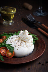 White homemade Burrata Italian cow milk cheese made of mozzarella and cream with soft texture seasoned with black pepper served with cherry tomatoes, arugula and live oil on dark brown wooden table