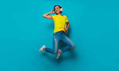 Fototapeta na wymiar Full-length photo of excited attractive shouting modern woman in yellow t-shirt and headphones with backpack while she jumping isolated on blue background. Happy study process