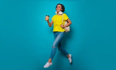 Fototapeta na wymiar Full-length photo of excited cute modern student girl in yellow t-shirt and headphones with backpack while she jumping isolated on blue background. Happy study process