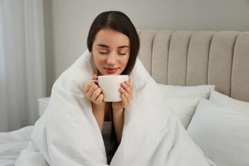 Beautiful young woman with cup of drink wrapped in blanket at home