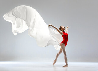 Ballerina in red ballet leotard dancing with white long cloth. She wears ballet pointe shoes. She dances in white studio