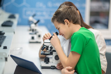 Boy looking through microscope near sitting teacher