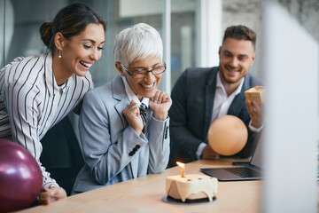 Happy senior CEO making a wish while blowing candle on Birthday cake during her surprise party in the office.