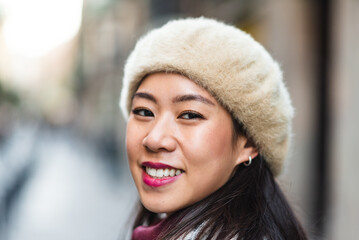 Cheerful Chinese woman with black hair in stylish beret looking at camera while standing on street against blurred background in city