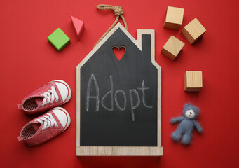 Small chalkboard with word ADOPT, toy bear, cubes and baby shoes on red background, flat lay