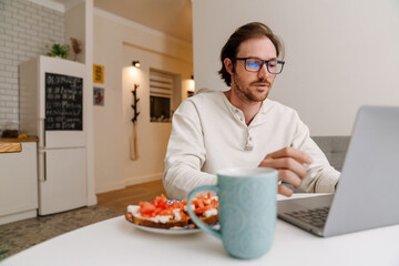 Ginger bristle man in eyeglasses working with laptop while having dinner