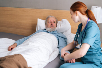 Fototapeta premium Female doctor measuring pulse on wrist to sick senior male patient lying on bed in hospital room. Nurse checking mature patient pulse at home. Elderly man getting medical consultation in hospice.