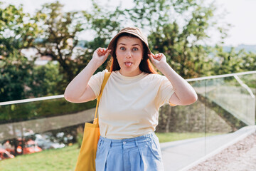Portrait of positive cheerful girl with tote bag over nature background, Summer time. Emotional stylish pretty young hipster woman sticking out her tongue