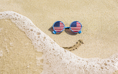 Fototapeta na wymiar Sunglasses with flag of USA on a sandy beach. Nearby is a sea lightning and a painted smile. The concept of a successful vacation in the resorts of USA.