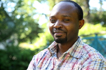 Naklejka na ściany i meble Young african man, portrait in his outdoor shirt, looking into the camera, outdoor in park, sunny atmosphere