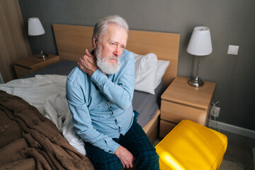 High-angle view of senior adult man warming up and massaging shoulder sitting on bed in morning...