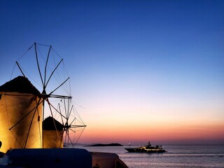 Mykonos windmill at sunset with yacht passing by - obrazy, fototapety, plakaty