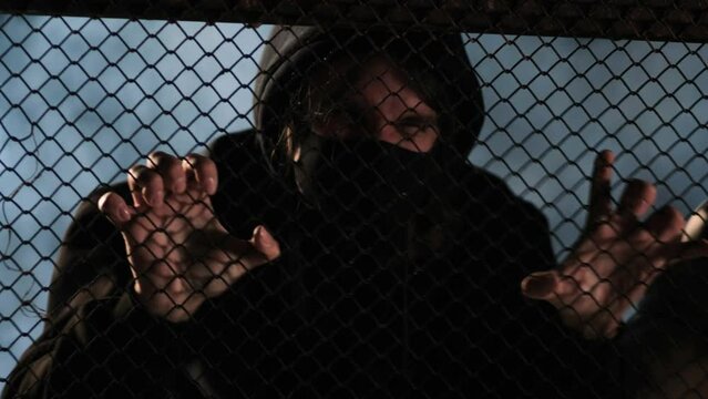 A violent aggressive bandit in a black mask is trying to break a mesh metal fence. A riot, an uprising