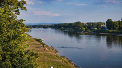 Obraz premium Panoramablick über die ruhig fließende Elbe mit grünen Elbwiesen und Uferlandschaft unter sommerlichem Himmel, Dresden, Sachsen, Deutschland