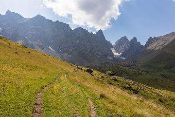 Fototapeta premium A panoramic view on the sharp mountain peaks of the Chaukhi massif in the Greater Caucasus Mountain Range in Georgia, Kazbegi Region. A hiking trail on a green alpine pasture. Georgian Dolomites.
