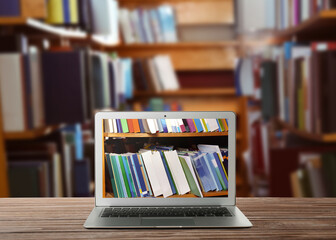 Online library. Modern laptop on wooden table and shelves with books indoors