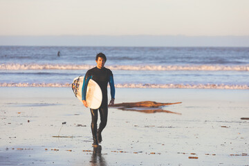 Young caucasian man walking on the beach with a surfboard.