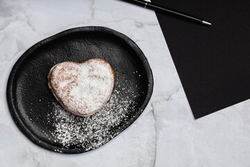 a heart-shaped donut on a plate, sprinkled with sugar, next to a piece of paper with a pen