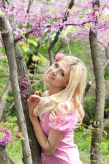 Cute young woman portrait with blonde hair posing in blooming pink flowers garden