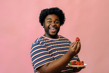 young happy african american man smiling looking at camera while eating strawberries in the studio over pink background