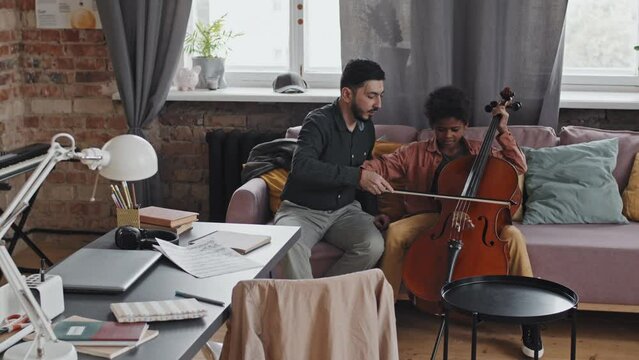 Wide shot of formally dressed Asian man sitting on couch in apartment at daytime, helping talented little Black boy playing double bass