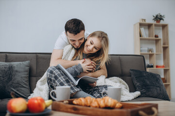 Couple in love sitting on the couch in the living room, wearing pajamas after getting up in the morning, enjoying the morning and having breakfast