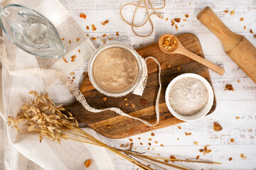 Homemade rye sourdough in glass jar with rye flour, rye ears, water, cereal grains and rolling pin on wooden background.