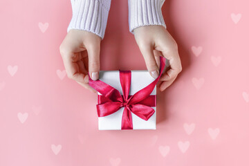 Female Hands in sweater holding gift in white wrapping paper on pink background with bokeh hearts. St. Valentines Day, love, tenderness, friendship and care concept. Cozy, festive, romantic wallpaper