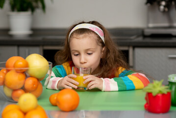 Fototapeta na wymiar A sweet little girl drink an orange fresh. She is dressed in multi-color t-shirt. She's in the kitchen at home. Healthy eating. Family. Fresh fruit.