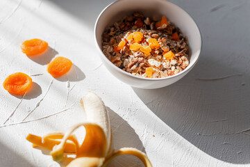 Healthy breakfast muesli with oatmeal, dried apricots and banana on a white table at sun lights in the morning