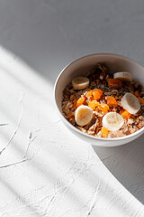 Healthy delicious raw breakfast muesli with oatmeal, dried apricots, banana and nuts in a white bowl on a white table at sunrise in morning