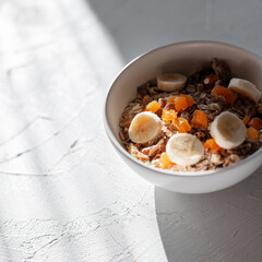Healthy tasty muesli with dried apricots, banana, nuts and dried fruits in a white bowl at sun lights in the morning. Fitness raw food and vegan diet