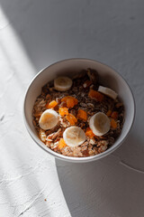 Delicious healthy food muesli with oatmeal, dried apricots, nuts and banana in a white bowl on a white table in sunlight. Vegan raw lactose free breakfast