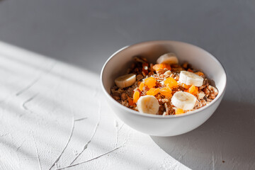 Delicious healthy breakfast muesli with natural oatmeal, dried apricots, nuts and bananas in a white bowl on a white table in the sun in the morning