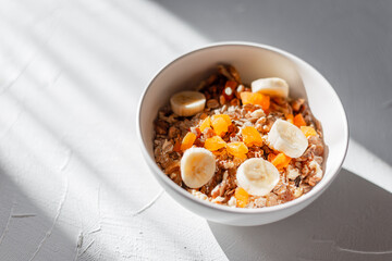 Delicious healthy breakfast exotic muesli with dried apricots, bananas, nuts and dried fruits in a white bowl on a white background in sunlight. Fitness raw food