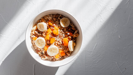 Delicious healthy breakfast muesli with oatmeal, dried apricots, walnuts and banana on a white table at sunrise in the morning