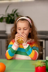 A sweet little girl drink an orange fresh. She is dressed in multi-color t-shirt. She's in the kitchen at home. Healthy eating. Family. Fresh fruit.