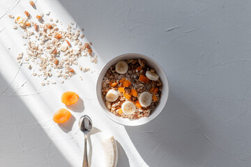 Healthy breakfast muesli with oatmeal, walnuts, dried apricots and banana on a white table at sun lights in the morning