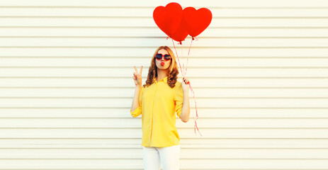 Portrait of beautiful young woman with red heart shaped balloon blowing her lips sending sweet air kiss wearing sunglasses on white background