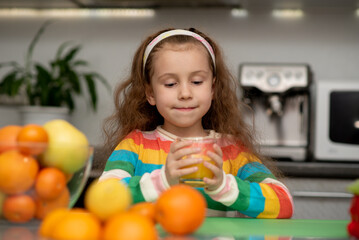 Fototapeta na wymiar A sweet little girl drink an orange fresh. She is dressed in multi-color t-shirt. She's in the kitchen at home. Healthy eating. Family. Fresh fruit.