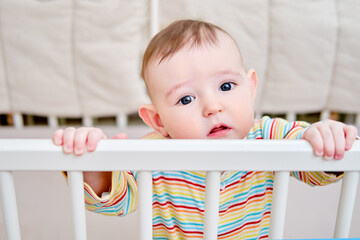 A sad baby in a crib, holding onto the handrails. Funny child in bed, age six months