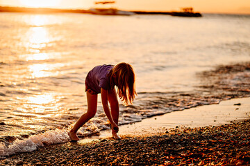 at sunset. kid girl walks along seashore, collects stones and throws into water