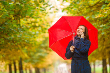 A young pretty woman walks down the alley with a red umbrella and talks on a mobile phone.