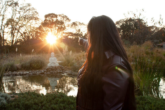 Woman dressed in purple leather jacket pointing her hand towards a golden sun that illuminates the buddha at sunset